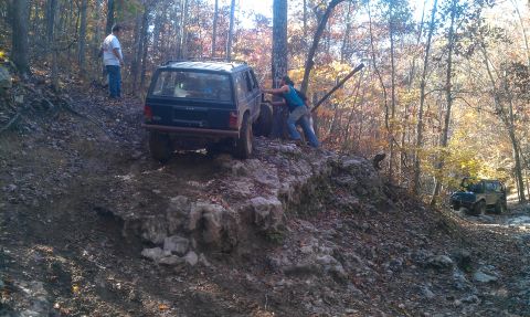 1987 Ford Bronco - Two off-road vehicles navigate a challenging, rocky trail in a wooded area during autumn.