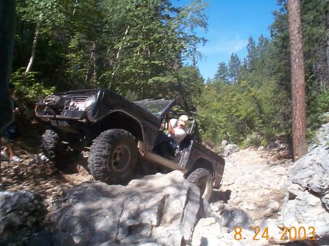 1977 Ford Bronco - A driver maneuvers an off-road vehicle over large rocks on a steep, wooded trail.