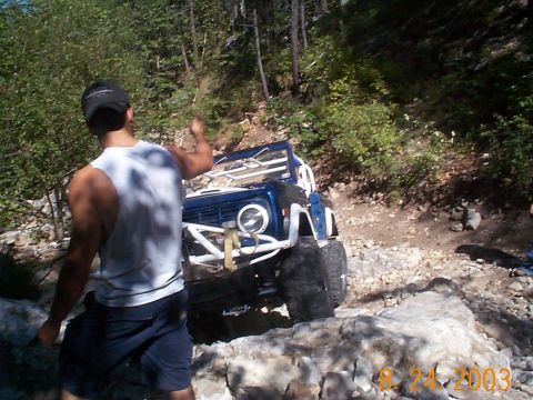 1977 Ford Bronco - A blue 1977 Ford Bronco navigates a rocky trail, equipped with a roll cage for off-road adventures.