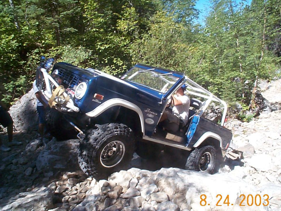 Blue Ford Bronco climbing over rocks on a trail. Driver is visible inside
