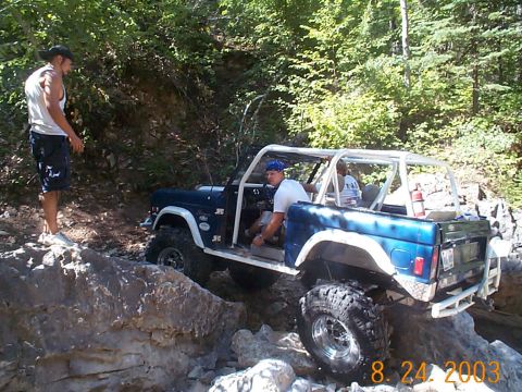 1977 Ford Bronco - A blue 1977 Ford Bronco navigates a challenging rocky trail with occupants inside and a person standing nearby.