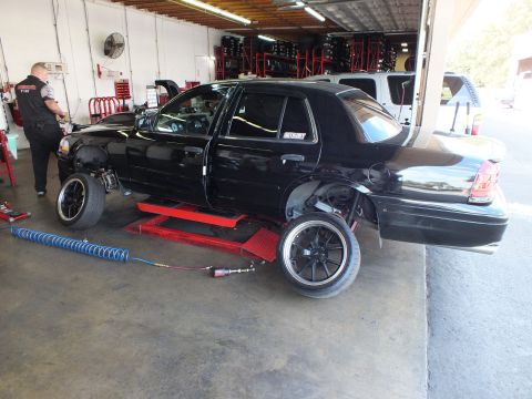 2003 Ford Crown Victoria - Black four-door sedan on a low-rise shop lift; a technician works at the front in a wheel-lined garage.