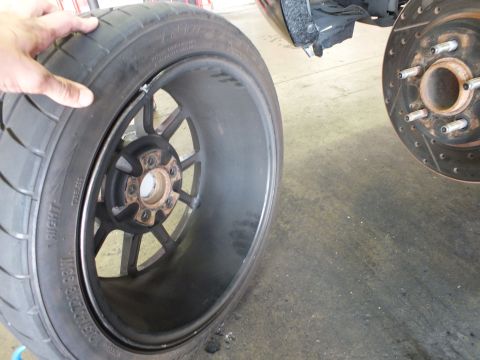 2003 Ford Crown Victoria - Black multi-spoke wheel held up in a shop; tire sidewall and a rusted brake hub visible behind.