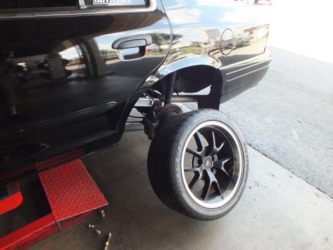 2003 Ford Crown Victoria - Rear corner of a black sedan raised on a lift; the wheel droops on its suspension above the floor.