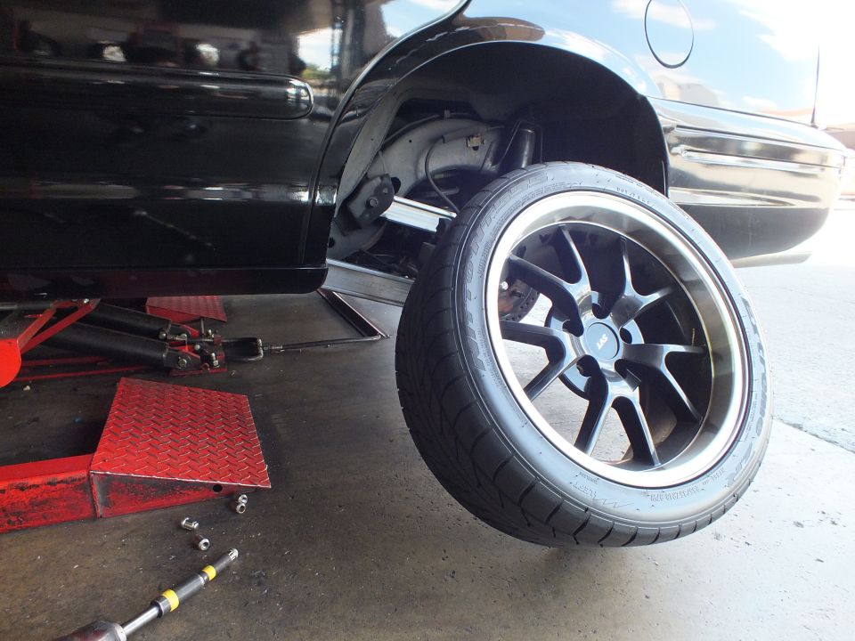 Black sedan raised on a lift showing rear suspension and black wheel with polished lip