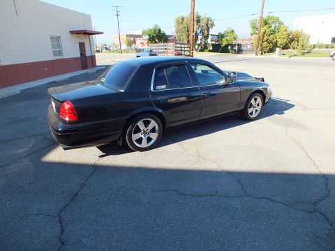 2003 Ford Crown Victoria - Lowered black four-door sedan on five-spoke wheels parked in a cracked asphalt lot near buildings.