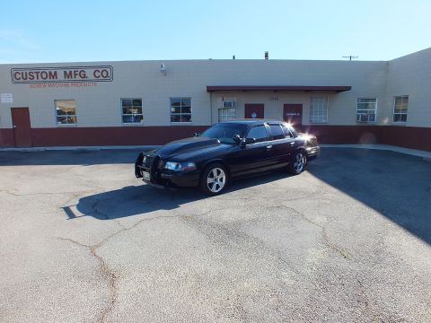2003 Ford Crown Victoria - Black sedan with push bar and chrome wheels sits in an empty lot fronting Custom Mfg. Co.