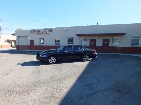 2003 Ford Crown Victoria - black push-bar Ford sits in a partly shaded parking lot fronting industrial buildings.