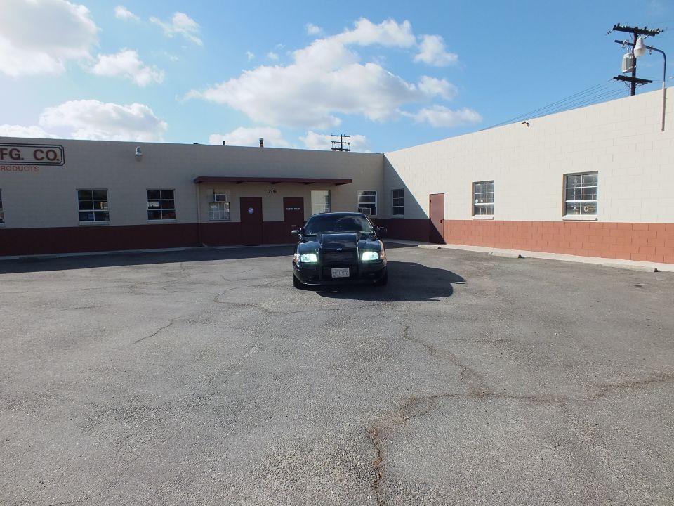 Black sedan with headlights on facing camera across wide industrial parking lot