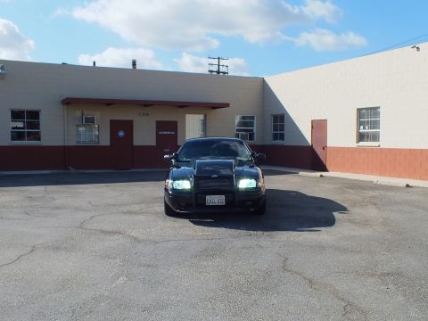 2003 Ford Crown Victoria - Frontal shot of the black sedan showing raised hood scoop and lit headlights near a warehouse.