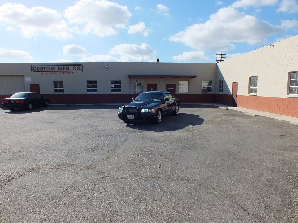 Black Ford sedan with headlights on parked beside Custom Mfg. Co. building