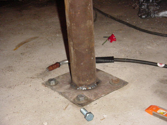 Welding torch attached to a metal pole base with bolts on a concrete floor. A small red and white object is nearby