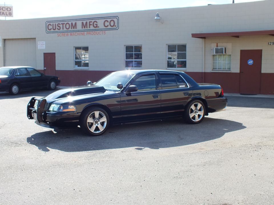 Black Crown Victoria with push bar and hood scoop fronting Custom Mfg. Co. building