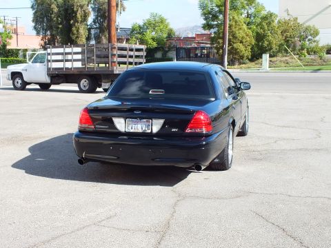 2003 Ford Crown Victoria - Tail end of a blacked-out full-size sedan in a lot, dual exhaust tips, EVIL VIC plate, work truck parked nearby.