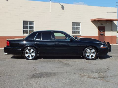 2003 Ford Crown Victoria - Sunlit side view of a black full-size sedan parked against a two-tone industrial wall.