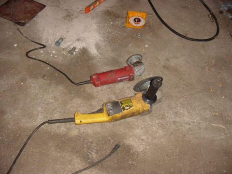1992 Ford Bronco - Two angle grinders, one red and one yellow, lie on a concrete floor next to some bolts and a level.