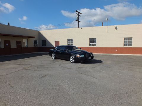 2003 Ford Crown Victoria - Black full-size sedan parked diagonally in a large empty industrial lot, headlights toward camera.