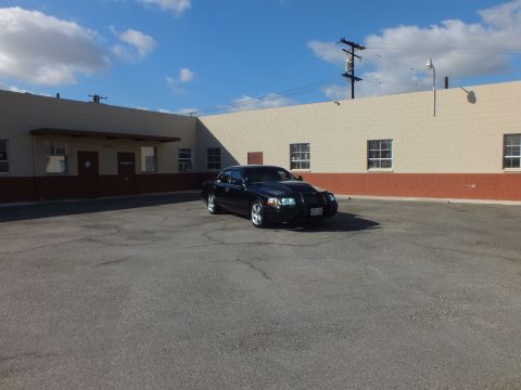 2003 Ford Crown Victoria - Front three-quarter of a black sedan with lights on, parked in a wide cracked parking lot.