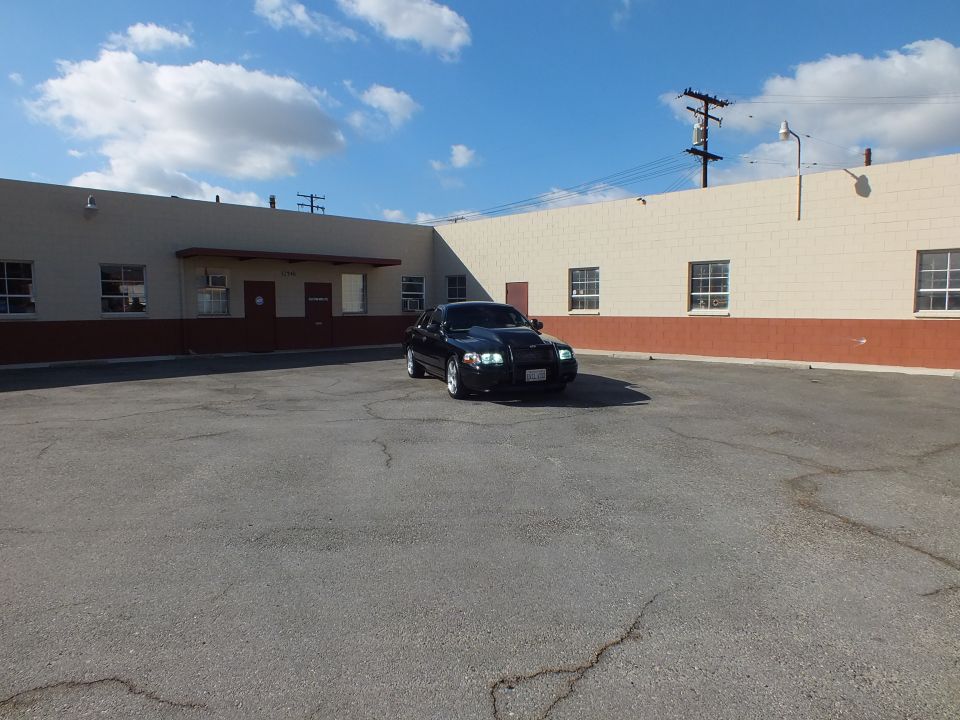 Black full-size sedan parked distant in empty lot between tan warehouse buildings