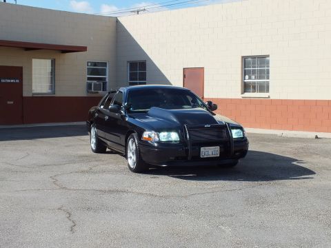 2003 Ford Crown Victoria - Black full-size sedan fitted with a brush guard, lights illuminated, parked by a tan industrial unit.