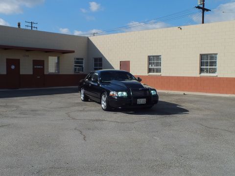 2003 Ford Crown Victoria - Front quarter of a black sedan with lit headlights in an industrial lot under scattered clouds.