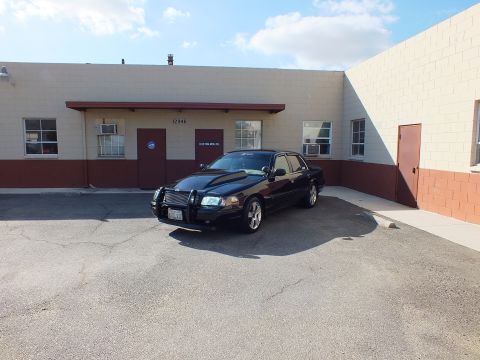 2003 Ford Crown Victoria - Black sedan with brush guard parked at an angle near a Custom Mfg Co warehouse entrance.