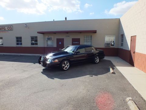 2003 Ford Crown Victoria - Black full-size sedan with brush guard parked beside an industrial building marked MFG CO.