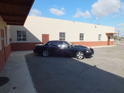 2003 Ford Crown Victoria - Rear-leaning profile of a black full-size sedan parked along a tan building wall.