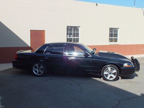 2003 Ford Crown Victoria - Side profile of a black full-size sedan, sun reflecting off the door, parked by a tan block wall.