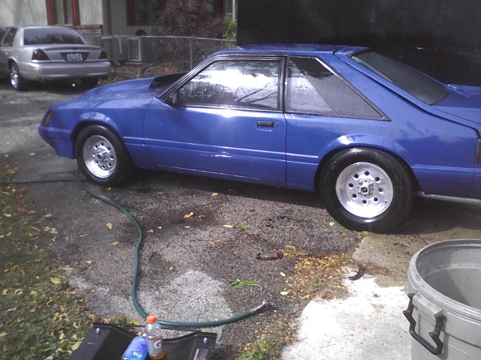 Blue coupe with aftermarket wheels and a hood scoop, parked on a wet driveway