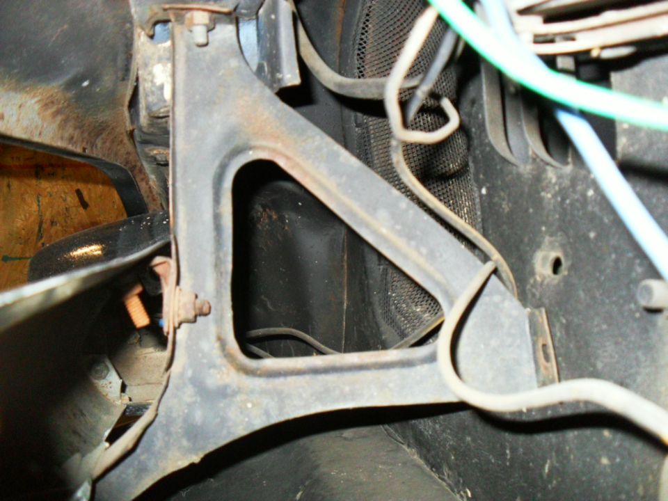Black triangular metal brace with surface rust, wiring, and latch hardware under the front end of a vintage Ford