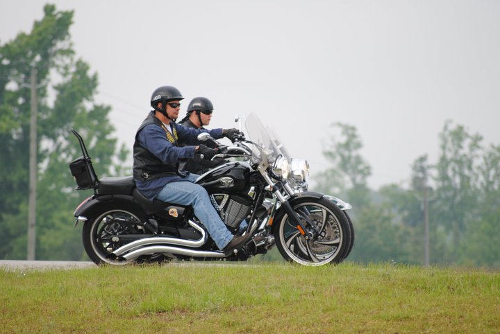 Two guys riding a black motorcycle on a road with grass and trees
