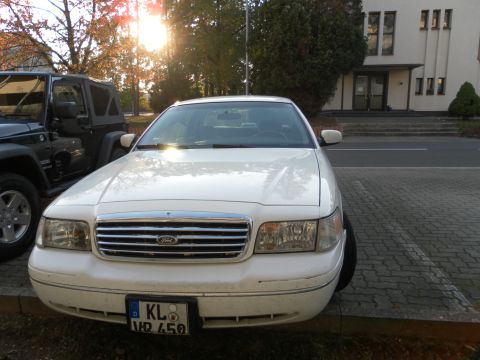 1998 Ford Crown Victoria - A white 1998 Ford Crown Victoria is parked in a lot next to a dark Jeep, with a building in the background.