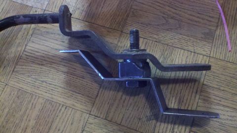 1978 Ford Bronco - Close-up of a metal bracket assembly, likely a vehicle part, resting on a wooden floor. Text from a watermark is visible.