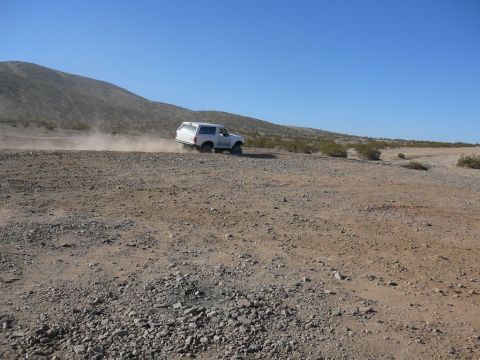 1988 Ford Bronco - A white 1988 Ford Bronco kicks up dust as it drives along a barren dirt road under a clear blue sky.