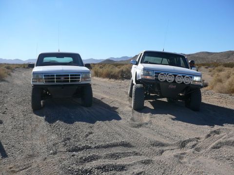 1988 Ford Bronco - Two white Ford trucks, likely from the early 1990s, are seen off-roading on a dirt path.