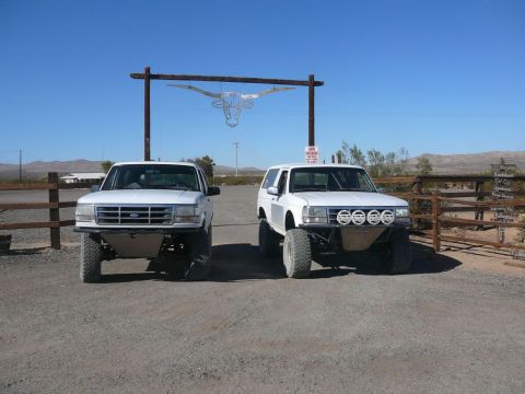1988 Ford Bronco - Two modified Ford Broncos are parked in front of a ranch entrance under a steer skull decoration.