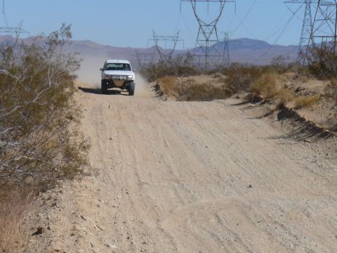 1988 Ford Bronco - A white off-road truck drives on a dusty desert road, with power lines visible in the background.