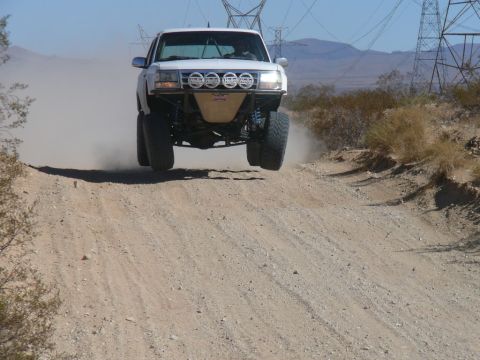 1988 Ford Bronco - A white off-road truck takes flight over a dirt hill, kicking up dust in a desert landscape.