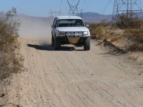 1988 Ford Bronco - White SUV with off-road modifications navigates a dusty desert track.