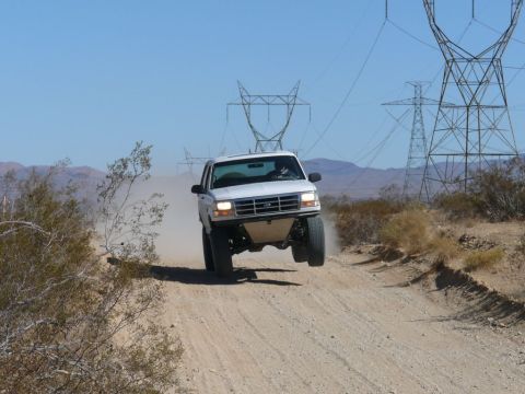 1988 Ford Bronco - A white Ford Bronco jumps over a dirt road in a desert landscape, with power lines in the background.