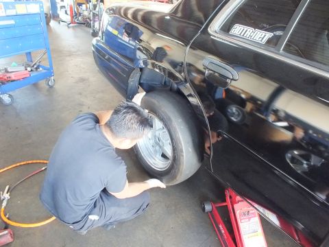 2003 Ford Crown Victoria - Worker crouches at the rear wheel well of a black sedan, fitting a drag tire on an aluminum wheel in a garage.