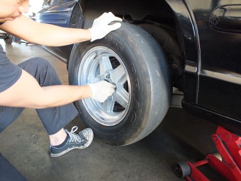 2003 Ford Crown Victoria - Kneeling worker holds an aluminum wheel and slick in place at the rear of a black car raised on a jack.