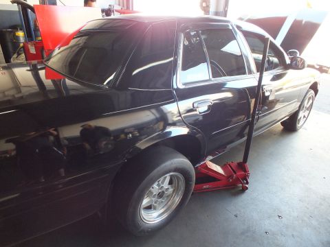 2003 Ford Crown Victoria - Black four-door sedan sits on a red floor jack in a garage with a polished drag wheel mounted at the rear.