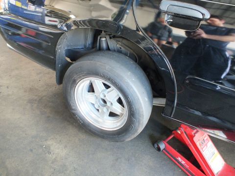 2003 Ford Crown Victoria - Wide drag tire on an aluminum wheel sits inside the rear wheel arch of a glossy black sedan in a shop.