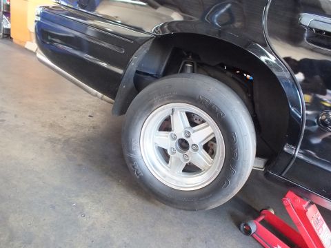 2003 Ford Crown Victoria - wide drag slick on a five-spoke aluminum wheel sits at the rear corner of a black car raised on a jack.
