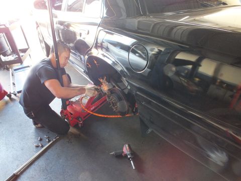 2003 Ford Crown Victoria - Worker uses a grinder on the exposed rear brake of a black sedan, throwing sparks with the wheel removed.