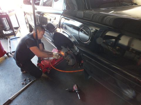 2003 Ford Crown Victoria - Kneeling worker grinds the rear brake area of a black car, with an impact wrench and lug nuts on the floor.