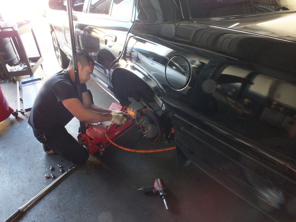Technician grinding exposed rear brake drum of black sedan on red jack, impact tools on floor