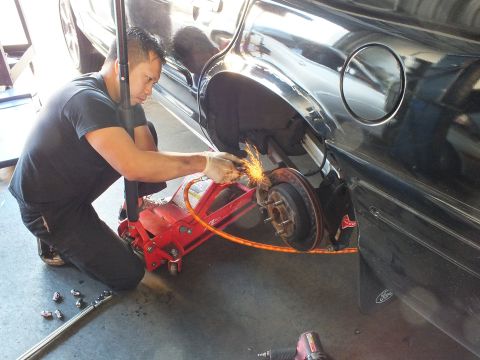 2003 Ford Crown Victoria - Technician throws sparks grinding the rear brake of a jacked black sedan, drill and lug nuts on the floor.
